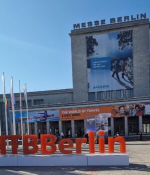 Grote rode letters spellen "#ITBBerlin" voor de Messe Berlin, een congrescentrum dat rijk is aan geschiedenis. Vlaggen omzomen het plein met spandoeken die reizen promoten, waaronder een voor Albanië. Het grijze gebouw staat hoog onder een helderblauwe hemel. | Echt Ierland Vakanties