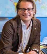 Een glimlachende persoon met een bril en lang haar, gekleed in een bruin jasje en lichtblauw shirt, zit aan een bureau met zijn handen op een smartphone. Achter hen suggereert een kleurrijke wereldkaart hun enthousiasme voor een aanstaande rondreis Ierland. | Echt Ierland Vakanties