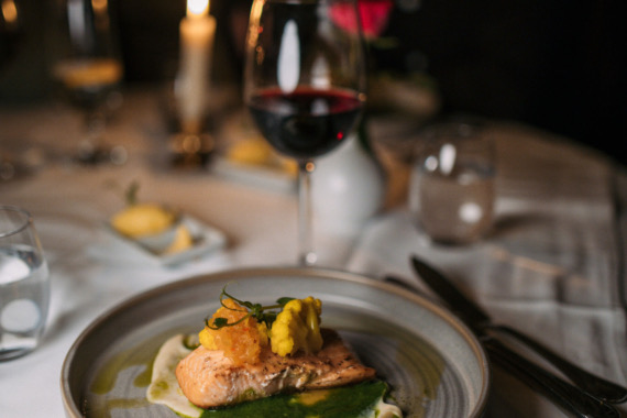 Een bord met gastronomisch eten met een gegrilde filet, gegarneerd met kruiden en decoratieve stukken, besprenkeld met groene saus, roept de elegantie op van een exclusieve eetervaring. Vergezeld van een glas rode wijn en geplaatst te midden van de charme die doet denken aan een exquise rondreis Ierland. | Echt Ierland Vakanties