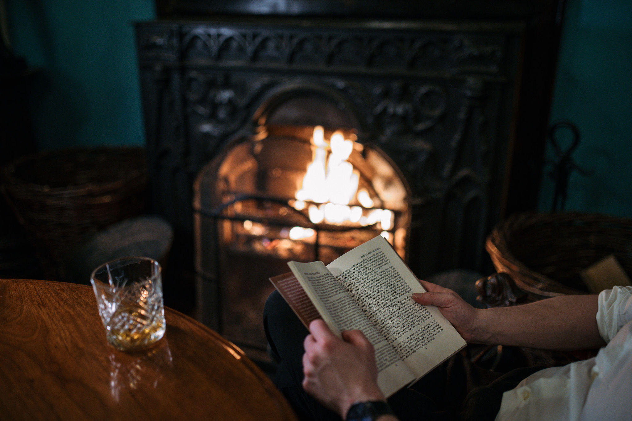 Een persoon zit bij een brandende open haard met een open boek in de hand. Naast hen staat een glas whisky op een houten tafel. De gezellige herfstavond straalt warmte uit en roept de charme van Ierland op als het haardlicht over hun handen en de bladzijden van het boek danst. | Echt Ierland Vakanties