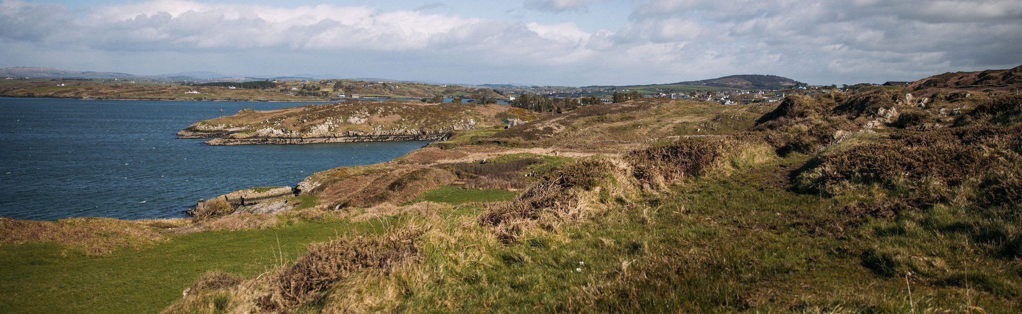 Een schilderachtig kustlandschap in Ierland met glooiende heuvels bedekt met gras en stukken heide. De zee is zichtbaar aan de linkerkant, die de horizon raakt. De lucht is overwegend helder met verspreide wolken, perfect voor een rondreis Ierland, aangezien kleine heuvels en verre formaties diepte toevoegen aan het pittoreske uitzicht. | Echt Ierland Vakanties