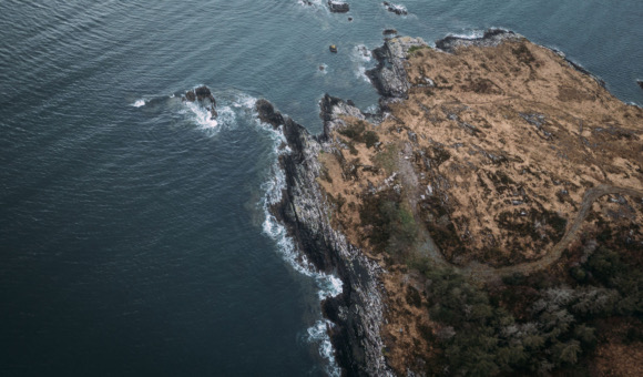 Luchtfoto van een ruige kustlijn in Ierland, met grillige kliffen en rotspartijen die zich uitstrekken tot in de donkerblauwe zee. Golven beuken zachtjes tegen de rotsen. Het land is bedekt met droog gras en stukken groen, wat een verbluffend contrast creëert: een perfect tafereel voor een rondreis Ierland. | Echt Ierland Vakanties