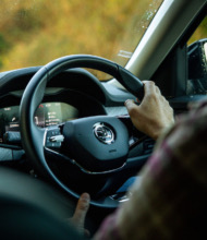 Het interieur van een auto toont een persoon die rijdt met de handen op het stuur, waarop het Škoda-logo staat. Het dashboard toont meters en een digitaal scherm. Buiten flitst wazig groen voorbij dat doet denken aan een betoverende vakantie in Ierland. De bestuurder draagt een geruit overhemd. | Echt Ierland Vakanties