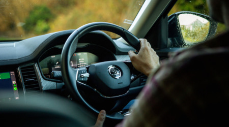 Het interieur van een auto toont een persoon die rijdt met de handen op het stuur, waarop het Škoda-logo staat. Het dashboard toont meters en een digitaal scherm. Buiten flitst wazig groen voorbij dat doet denken aan een betoverende vakantie in Ierland. De bestuurder draagt een geruit overhemd. | Echt Ierland Vakanties