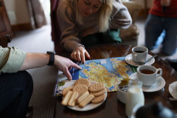 Een groep mensen verzamelde zich rond een salontafel met een kaart, enthousiast pratend over hun aankomende rondreis Ierland. Kopjes koffie, een fles melk en diverse koekjes liggen verspreid op de tafel. De gezellige binnenomgeving heeft een vloerbedekking en gestoffeerde meubels, wat bijdraagt aan de voorpret van hun Ierse avontuur. | Echt Ierland Vakanties
