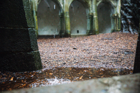 Natte grond in een stenen binnenplaats, bezaaid met gevallen bruine bladeren. Regenwater creëert rimpelingen in plassen, die de serene charme van een rondreis Ierland omsluiten. De achtergrond bestaat uit gebogen deuropeningen en een stenen muur, die de vredige sfeer oproepen die kenmerkend is voor een vakantie in Ierland. | Echt Ierland Vakanties