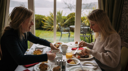 Twee vrouwen dineren aan een tafel bij een groot raam met uitzicht op een weelderige tuin en het water, wat doet denken aan een schilderachtige vakantie in Ierland. Ze genieten van scones, sinaasappelsap en conserven in een gezellige, informele omgeving die wordt verlicht door natuurlijk licht. | Echt Ierland Vakanties