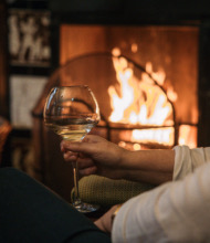 In een gezellige setting die doet denken aan een vakantie in Ierland, houdt een persoon een glas witte wijn vast, zittend voor een brullende open haard. De warme gloed versterkt de intieme sfeer, met een gevlochten mand die gedeeltelijk zichtbaar is aan de zijkant. Het is alsof je geniet van de rustige charmes van een Ierland-tour vanuit huis. | Echt Ierland Vakanties