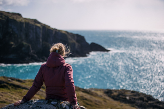 Een persoon in een bordeauxrood jasje zit op een rots, uitkijkend over de uitgestrekte, sprankelende blauwe oceaan van Ierland. In de verte zijn kliffen te zien onder een gedeeltelijk bewolkte lucht, die rust en een diepe verbinding met de natuur oproepen - een ideaal moment voor degenen die op rondreis Ierland zijn. | Echt Ierland Vakanties