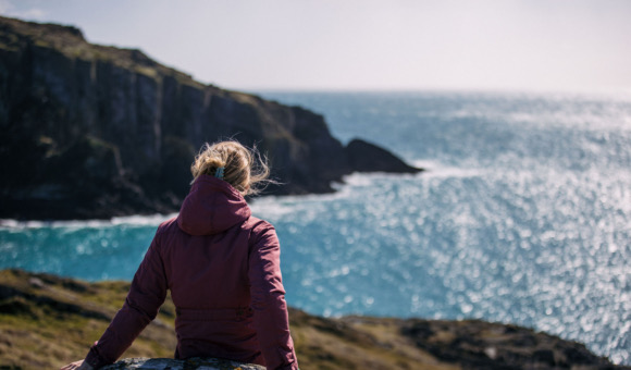 Een persoon in een bordeauxrood jasje zit op een rots, uitkijkend over de uitgestrekte, sprankelende blauwe oceaan van Ierland. In de verte zijn kliffen te zien onder een gedeeltelijk bewolkte lucht, die rust en een diepe verbinding met de natuur oproepen - een ideaal moment voor degenen die op rondreis Ierland zijn. | Echt Ierland Vakanties