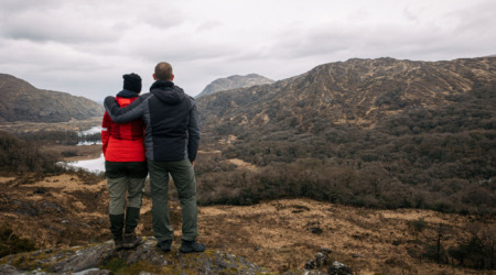 Twee mensen staan op een rotsachtige richel, uitkijkend over een uitgestrekt, bergachtig Iers landschap. De een draagt een rood jasje en de ander een donker jasje. Hun armen zijn om elkaar heen geslagen, genietend van de schoonheid van de rondreis Ierland met glooiende heuvels, bewolkte luchten en een klein waterlichaam eronder. | Echt Ierland Vakanties