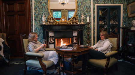 Twee vrouwen zitten in een vintage woonkamer, verwikkeld in gesprek bij een brullende open haard. De ruimte, die doet denken aan gezellige Ierland vakanties, heeft sierlijk behang en een grote ronde spiegel boven de schouw. Hun warme dialoog ontvouwt zich alsof ze op een schilderachtige rondreis Ierland zijn. | Echt Ierland Vakanties