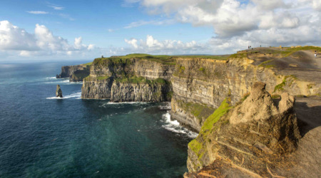 Kliffen die zich uitstrekken langs een oceaankustlijn in de pittoreske setting van een vakantie Ierland, met steile, gelaagde rotsformaties en verspreid groen. De voorgrond toont een ruig pad, terwijl de achtergrond een uitgestrekte zee onder een gedeeltelijk bewolkte lucht laat zien. Een paar mensen bovenop de kliffen benadrukken hun omvang, perfect voor degenen die een avontuurlijke rondreis Ierland maken. | Echt Ierland Vakanties