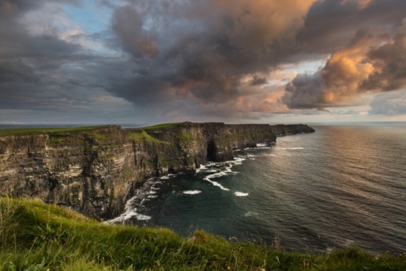 Ervaar een dramatisch kustlandschap tijdens uw vakantie in Ierland bij de Cliffs of Moher. Rotsachtige kliffen met groene toppen rijzen scherp uit boven de oceaan, waar golven beuken onder een lucht vol humeurige wolken en flarden zonsonderganglicht. Weelderig gras maakt dit pittoreske tafereel compleet. | Echt Ierland Vakanties