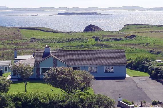 Een blauw, gelijkvloers huis met een donker betegeld dak is omgeven door groene gazons en bomen, perfect voor degenen die dromen van een rondreis door Ierland. Een brede oprijlaan leidt naar de ingang. Op de achtergrond strekt een graslandschap zich uit tot een glinsterend waterlichaam en verre eilanden onder een heldere hemel. | Echt Ierland Vakanties