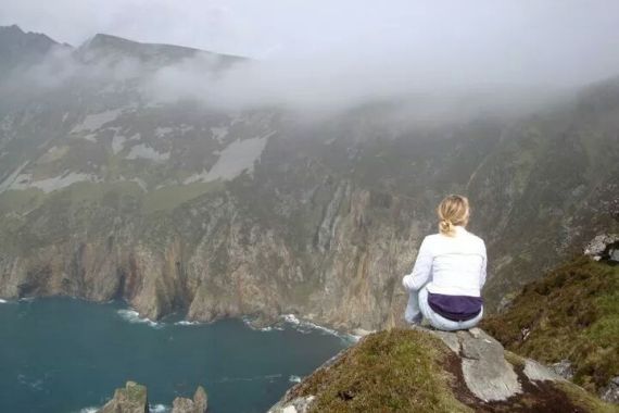 Een persoon met blond haar, in een lichte jas en spijkerbroek, zit op een rotsachtige heuvel met uitzicht op dramatische zeekliffen - een sereen tafereel dat deel zou kunnen uitmaken van een 10-daagse fly-drive vakantie Ierland, met mist die de smaragdgroene toppen in de verte en de blauwgroene zee eronder verhult. | Echt Ierland Vakanties