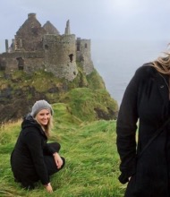 Een vrouw in een zwarte jas en grijze muts staat lachend op grasachtige kliffen met de ruïnes van Dunluce Castle op de achtergrond, en legt de essentie van haar vakantie in Ierland vast. Een andere afbeelding toont haar gehurkt, terwijl de bewolkte lucht en de oceaan het dramatische, ruige landschap benadrukken dat kenmerkend is voor vakanties in Ierland. | Echt Ierland Vakanties