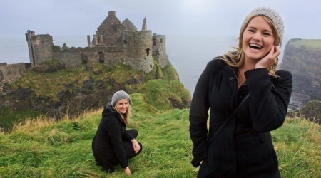 Een vrouw in een zwarte jas en grijze muts staat lachend op grasachtige kliffen met de ruïnes van Dunluce Castle op de achtergrond, en legt de essentie van haar vakantie in Ierland vast. Een andere afbeelding toont haar gehurkt, terwijl de bewolkte lucht en de oceaan het dramatische, ruige landschap benadrukken dat kenmerkend is voor vakanties in Ierland. | Echt Ierland Vakanties