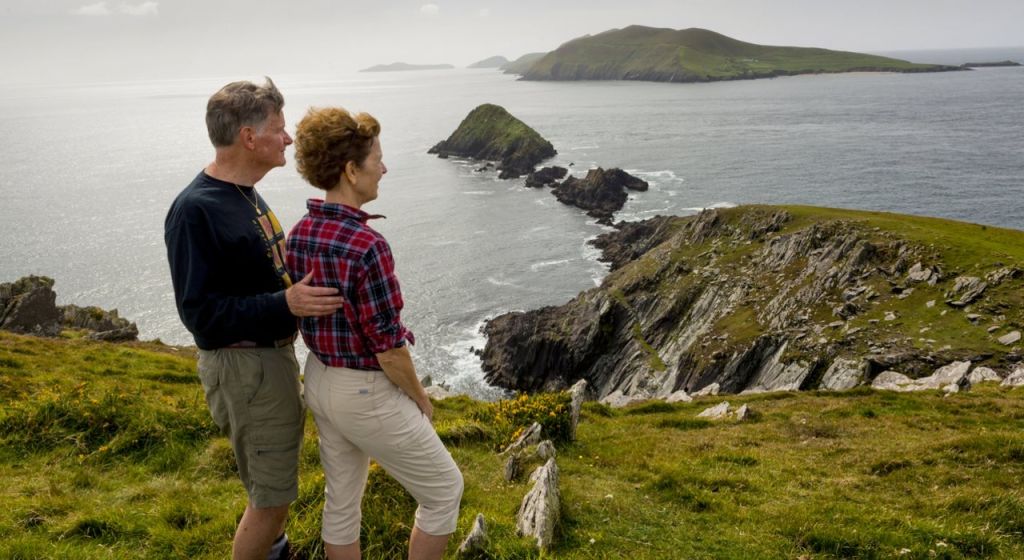 Een stel staat op een met gras begroeide klif tijdens hun rondreis Ierland, met uitzicht op een ruige kustlijn en verre eilanden onder een bewolkte lucht. De man draagt een zwart shirt, terwijl de vrouw een rood geruit shirt draagt. Oceaan golven beuken tegen de kust, met groene heuvels op de achtergrond. | Echt Ierland Vakanties