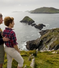 Een stel staat op een met gras begroeide klif tijdens hun rondreis door Ierland, met uitzicht op een ruige kustlijn. De man heeft zijn arm om de vrouw heen terwijl ze naar verre bergen en een kalme zee onder een bewolkte lucht staren. Het landschap bestaat uit steile rotswanden die zich uitstrekken tot in de oceaan met weelderige groene heuvels erachter. | Echt Ierland Vakanties