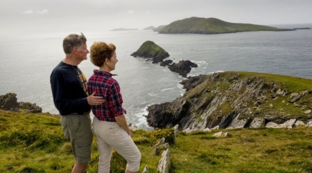 Een stel staat op een met gras begroeide klif tijdens hun rondreis door Ierland, met uitzicht op een ruige kustlijn. De man heeft zijn arm om de vrouw heen terwijl ze naar verre bergen en een kalme zee onder een bewolkte lucht staren. Het landschap bestaat uit steile rotswanden die zich uitstrekken tot in de oceaan met weelderige groene heuvels erachter. | Echt Ierland Vakanties