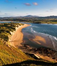 Luchtfoto van een kustlandschap in Ierland met een zandstrand dat langs de kust loopt. Links strekken zich grasduinen en groene velden uit landinwaarts. Golven klotsen zachtjes tegen de kust aan de rechterkant. In de verte maken glooiende heuvels en bergen onder een blauwe lucht met verspreide wolken dit perfecte vakantie Ierland-tafereel compleet. | Echt Ierland Vakanties