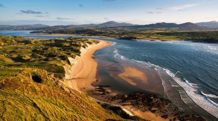 Luchtfoto van een kustlandschap in Ierland met een zandstrand dat langs de kust loopt. Links strekken zich grasduinen en groene velden uit landinwaarts. Golven klotsen zachtjes tegen de kust aan de rechterkant. In de verte maken glooiende heuvels en bergen onder een blauwe lucht met verspreide wolken dit perfecte vakantie Ierland-tafereel compleet. | Echt Ierland Vakanties