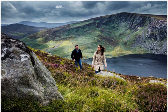 Een man en een vrouw wandelen een met gras begroeide, rotsachtige heuvel op met uitzicht op een meer en bergachtig landschap, wat de essentie van een vakantie Ierland belichaamt. De bewolkte lucht met dramatische wolken en paarse heide op de voorgrond geven kleur aan het ruige landschap, wat hun onvergetelijke ervaring nog beter maakt. | Echt Ierland Vakanties