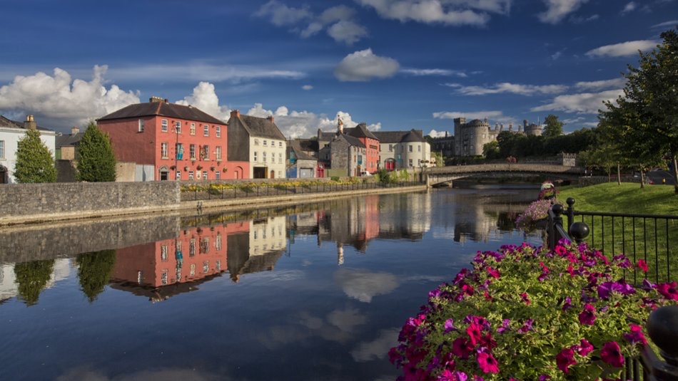 Schilderachtig uitzicht op een rivier met kleurrijke gebouwen en een stenen brug weerspiegeld in het kalme water tijdens een vakantie in Ierland. Een levendig bloemenperk met roze en paarse bloesems siert de voorgrond, terwijl een middeleeuws kasteel op de achtergrond staat onder een blauwe, gedeeltelijk bewolkte lucht. | Echt Ierland Vakanties