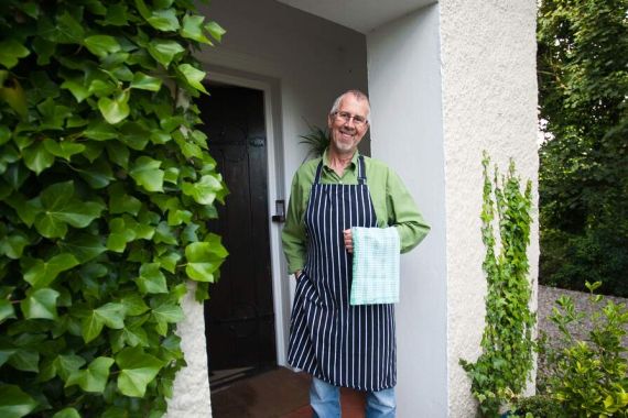 Een glimlachende man staat in een deuropening omringd door groene klimop, wat de charme van een vakantie in Ierland oproept. Hij draagt een groen shirt, een gestreept schort, jeans en witte sneakers, en houdt een geruite doek vast. De witte muur met textuur en het weelderige groen doen denken aan de pittoreske landschappen van reizen Irland. | Echt Ierland Vakanties