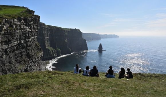 Een groep mensen zit op grasachtige kliffen, genietend van hun vakantie in Ierland en de adembenemende Cliffs of Moher bewonderend. De ruige kustlijn strekt zich uit met de oceaan eronder, terwijl een helderblauwe lucht en zonlicht dat op het water schittert een sereen en pittoresk tafereel vormen. | Echt Ierland Vakanties