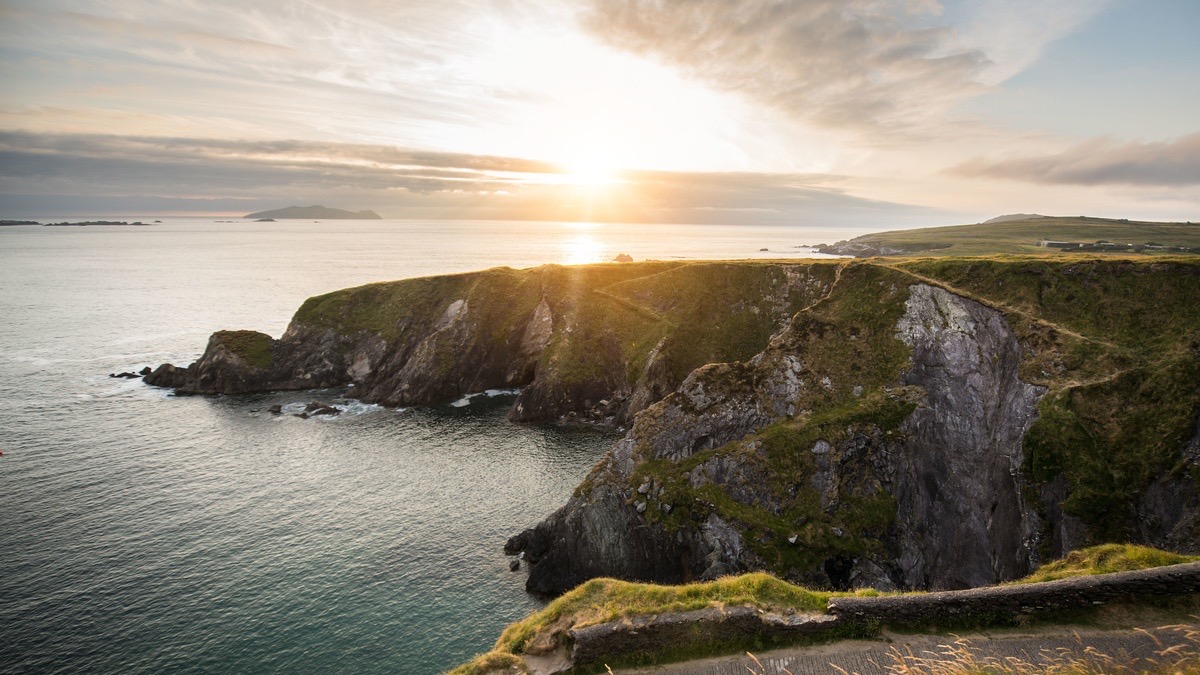 Zonsondergang boven ruige kliffen en kalme oceaanwateren schetst een pittoresk tafereel dat doet denken aan een rondreis Ierland. De zon werpt een warme gloed, die de rotsachtige kustlijn en de met gras begroeide kliftoppen benadrukt. Wolken weerspiegelen tinten oranje en roze, met een eiland in de verte dat de schilderachtige schoonheid versterkt. | Echt Ierland Vakanties