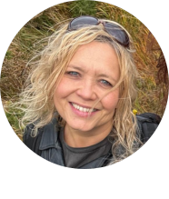 De vrouw, stralend lachend in haar zwarte jas en zonnebril op haar hoofd, staat te midden van het torenhoge gras en de planten, wat het gevoel van een buitenavontuur oproept. Haar blonde, krullende haar gloeit in de zon terwijl ze geniet van haar vrolijke en ontspannen rundreise Irland. | Echt Ierland Vakanties