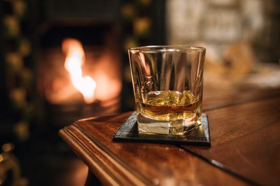 Een glas whisky op een onderzetter rust op een houten tafel, wat herinneringen oproept aan een ontspannende urlaub Ierland. Op de wazige achtergrond draagt een open haard met een warm, gloeiend vuur bij aan de gezellige ambiance, wat een intieme en uitnodigende sfeer suggereert. | Echt Ierland Vakanties