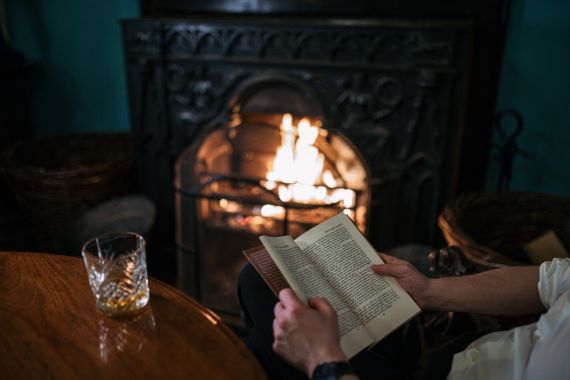 Een persoon zit bij een brandende open haard, met een open boek in de hand. Een glas whisky, dat doet denken aan die van een vakantie in Ierland, staat op een houten tafel op de voorgrond. Rieten manden flankeren de sierlijke zwarte open haard, wat een gezellige sfeer creëert tegen de achtergrond van de blauwgroene muur. | Echt Ierland Vakanties