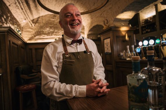 Een lachende barman met een witte baard, een vlinderdas en een schort, staat achter een houten bar. Het rustieke gewelfde plafond en de warme verlichting roepen de charme van een Ierse pub op, een perfecte plek om te ontspannen na uw "urlaub Irland", terwijl flessen sterke drank en taps de achtergrond vormen. | Echt Ierland Vakanties