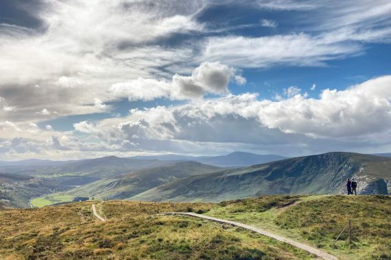Een uitgestrekt landschap toont een met gras begroeide heuveltop met een kronkelend pad dat in de verte leidt, perfect voor degenen die een urlaub in Ierland plannen. Twee mensen staan rechts en kijken uit over uitgestrekte groene valleien beneden. De dramatische lucht is gevuld met donzige wolken tegen een achtergrond van blauw, terwijl de heuvels zich uitstrekken tot aan de horizon. | Echt Ierland Vakanties