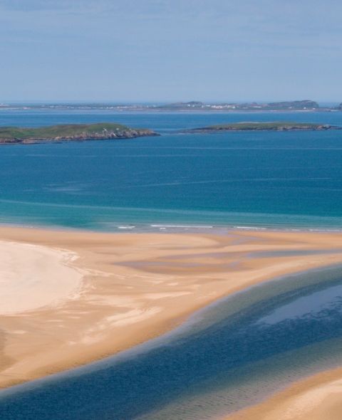 Luchtfoto van een kustlijn in Ierland waar gouden zandstranden de turquoise zee ontmoeten. Een smalle, donkerblauwe rivier slingert door het zand de oceaan in. In de verte stippelen groene, met gras begroeide eilanden de horizon onder een helderblauwe lucht, perfect voor uw nächste Urlaub Irland-avontuur. | Echt Ierland Vakanties
