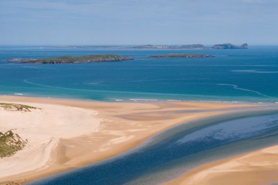 Luchtfoto van een kustlijn in Ierland waar gouden zandstranden de turquoise zee ontmoeten. Een smalle, donkerblauwe rivier slingert door het zand de oceaan in. In de verte stippelen groene, met gras begroeide eilanden de horizon onder een helderblauwe lucht, perfect voor uw nächste Urlaub Irland-avontuur. | Echt Ierland Vakanties