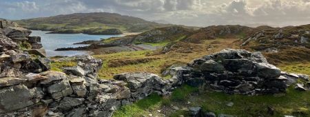 Uitzicht op een ruig kustlandschap met stenen ruïnes op de voorgrond, perfect voor degenen die op een "urlaub Irland" zijn. De lucht is gedeeltelijk bewolkt en zonlicht werpt zachte schaduwen op de heuvels en het grasachtige terrein. De kalme oceaan ontmoet de rotsachtige kustlijn, waardoor een serene atmosfeer ontstaat onder een blauwe lucht. | Echt Ierland Vakanties
