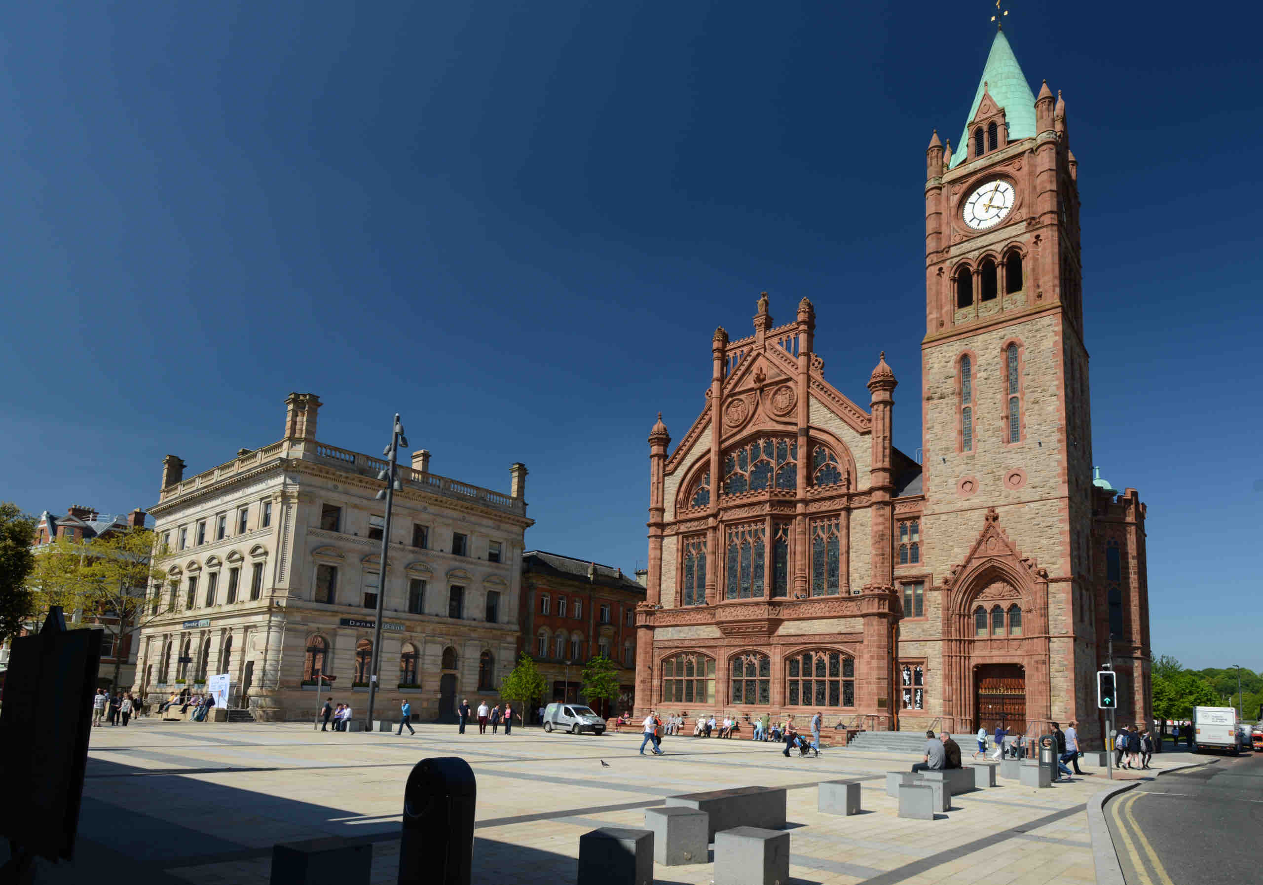 Een levendig stadsplein in Ierland met een historisch roodstenen gebouw met een hoge klokkentoren onder een helderblauwe lucht. Mensen slenteren ontspannen over het zonnige plein en vangen de essentie van een betoverende rondreis Ierland. Een elegant stenen gebouw draagt bij aan de architectonische charme. | Echt Ierland Vakanties