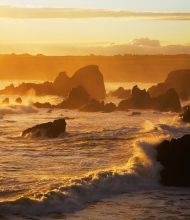 Een rotsachtige kustlijn bij zonsondergang in Ierland, met golven die tegen scherpe, donkere rotsen slaan. De lucht is goudkleurig met zachte wolken, die een warme gloed over de woelige zee werpen. De horizonlijn hint naar avontuurlijke rondreis Ierland-mogelijkheden, waarbij rust en beweging worden gecombineerd in dit betoverende tafereel. | Echt Ierland Vakanties