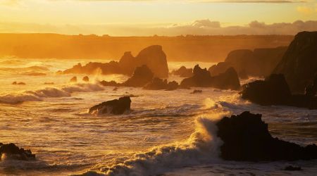Een rotsachtige kustlijn bij zonsondergang in Ierland, met golven die tegen scherpe, donkere rotsen slaan. De lucht is goudkleurig met zachte wolken, die een warme gloed over de woelige zee werpen. De horizonlijn hint naar avontuurlijke rondreis Ierland-mogelijkheden, waarbij rust en beweging worden gecombineerd in dit betoverende tafereel. | Echt Ierland Vakanties