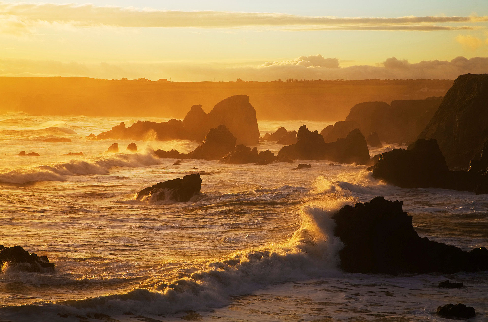 Een rotsachtige kustlijn bij zonsondergang in Ierland, met golven die tegen scherpe, donkere rotsen slaan. De lucht is goudkleurig met zachte wolken, die een warme gloed over de woelige zee werpen. De horizonlijn hint naar avontuurlijke rondreis Ierland-mogelijkheden, waarbij rust en beweging worden gecombineerd in dit betoverende tafereel. | Echt Ierland Vakanties
