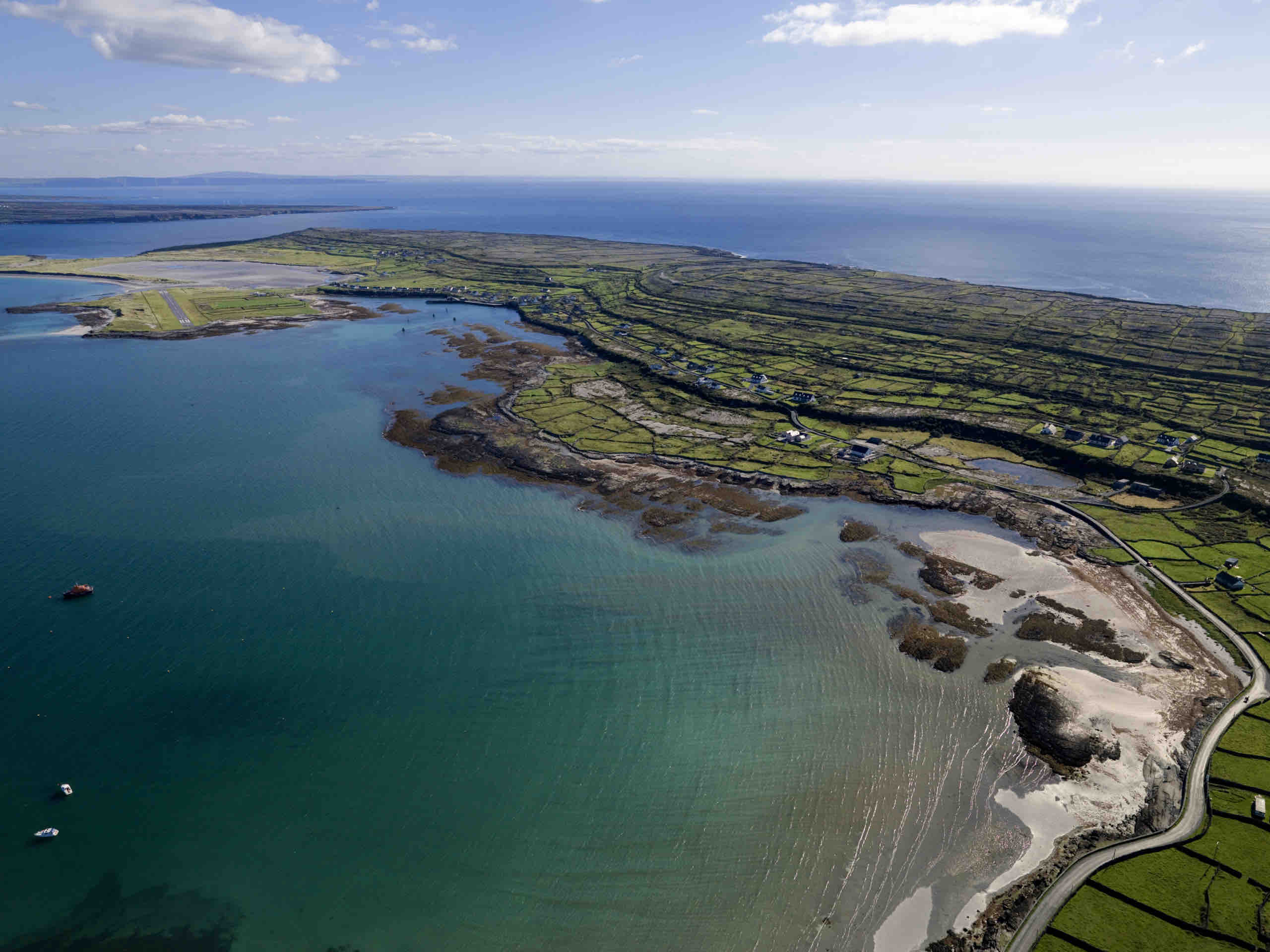 Luchtfoto van een kustlandschap in Ierland met een kustlijn met rotsachtige plekken en helder turquoise water. Het land is verdeeld in groene velden, omlijnd door stenen muren. Perfect voor een rondreis Ierland, een smalle weg loopt langs de kust onder een helderblauwe lucht met verspreide wolken erboven. | Echt Ierland Vakanties