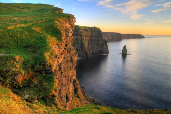 De afbeelding legt de Cliffs of Moher in Ierland vast bij zonsondergang, een must-see op elke rondreis door Ierland. Enorme kliffen storten zich in de kalme Atlantische Oceaan, met een oranje en roze lucht weerspiegeld in het water. Groen gras siert de kliftoppen, terwijl een eenzame zeestapel in de buurt oprijst. | Echt Ierland Vakanties