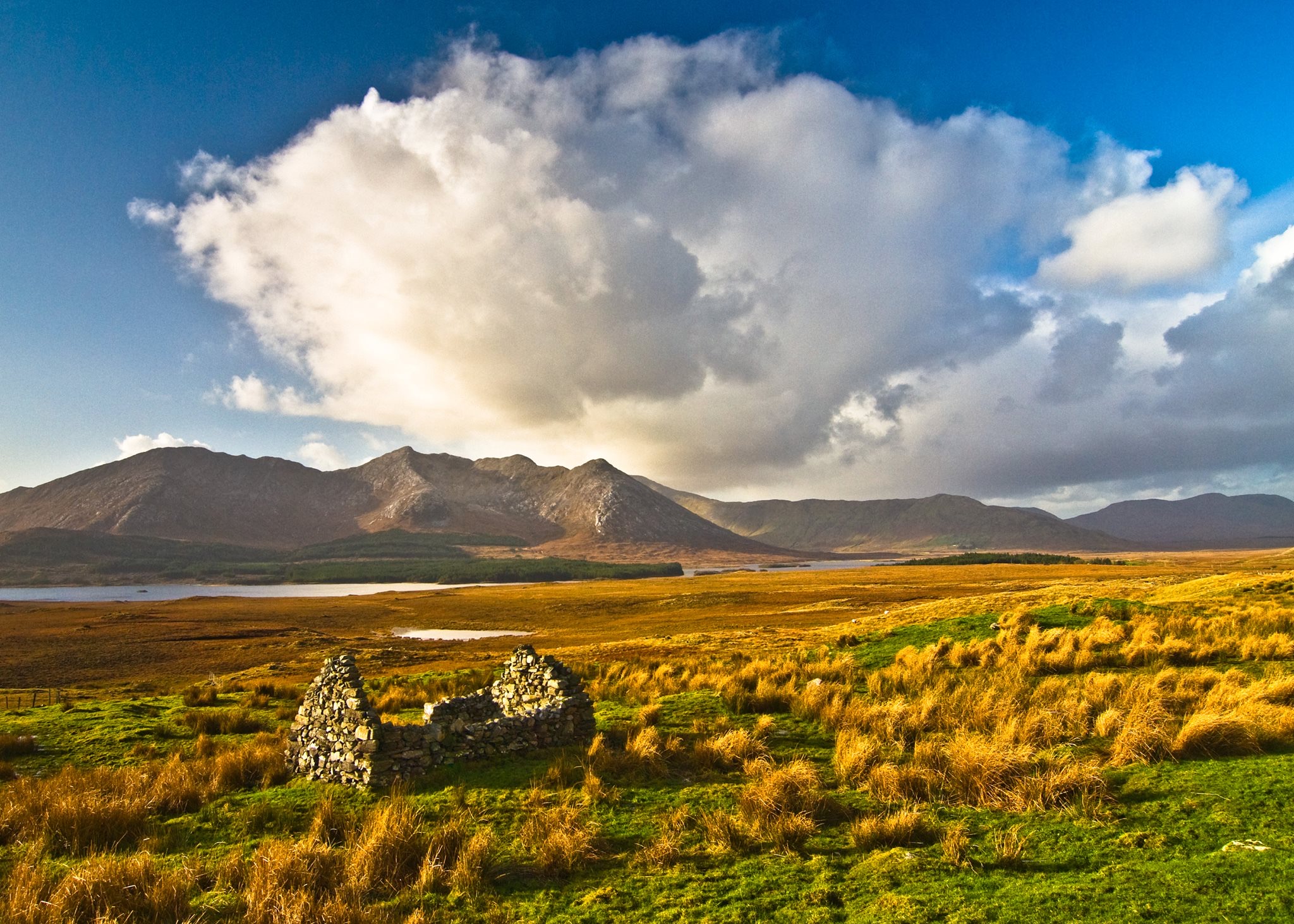 Te midden van het graslandschap met goudbruine tinten staat een stenen ruïne stil. Op de achtergrond is een uitgestrekte bergketen onder de helderblauwe lucht gedeeltelijk verhuld door donzige wolken. Een glinsterend meer draagt bij aan dit serene en uitgestrekte uitzicht, ideaal voor een rondreis Ierland of vakantie Ierland. | Echt Ierland Vakanties