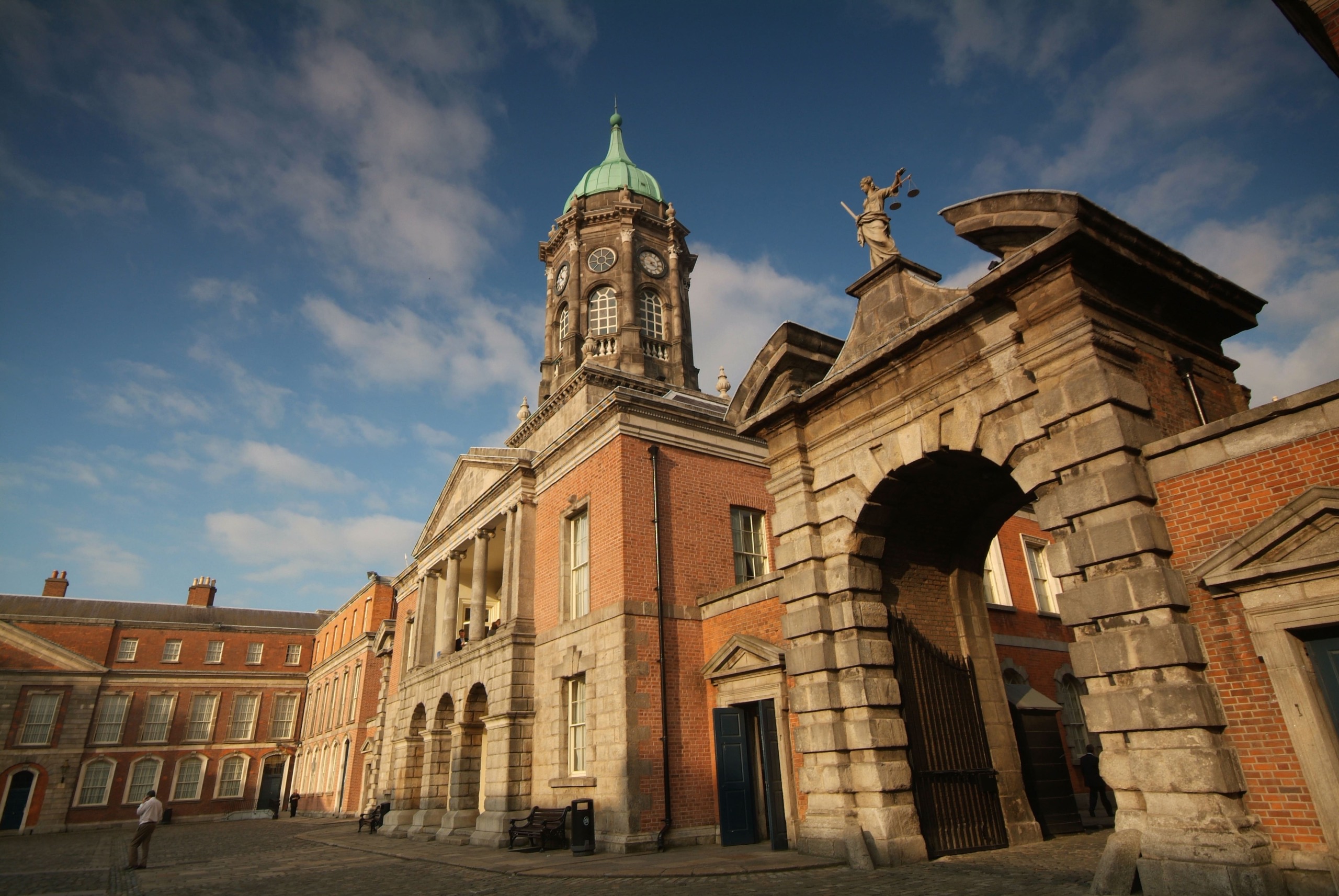Dit historische kasteelcomplex in Ierland, perfect voor een "Ierland vakantie", heeft een majestueuze klokkentoren, een gewelfde ingang en charmante bakstenen gebouwen. De overwegend heldere lucht en het warme zonlicht versterken de verbluffende architectuur met zijn zuilen en standbeelden, terwijl een eenzame figuur schaal toevoegt aan het tafereel. | Echt Ierland Vakanties