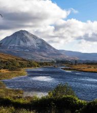 Ontdek de betoverende schoonheid van een schilderachtig landschap dat perfect is voor een vakantie in Ierland. Een kronkelende rivier geflankeerd door weelderig groen en een bladloze boom siert de voorgrond, terwijl een majestueuze berg oprijst tegen een door wolken gekuste lucht, schaduwen werpend op glooiende heuvels en gouden weiden. | Echt Ierland Vakanties