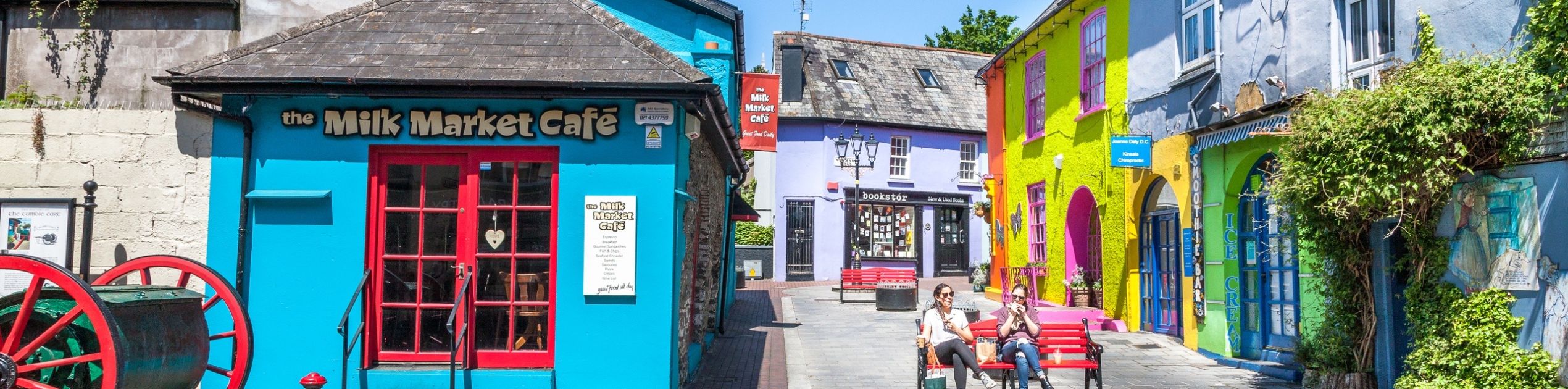 Kleurrijke straatscène die doet denken aan reizen Irland, met levendige gebouwen in tinten blauw, paars, geel en groen. Het "Milk Market Café" valt op als mensen op rode bankjes zitten. Een hoog grijs gebouw met het label "Grey Hound" doemt op in de achtergrond met een groot rood wiel aan de linkerkant onder een zonnige hemel. | Echt Ierland Vakanties